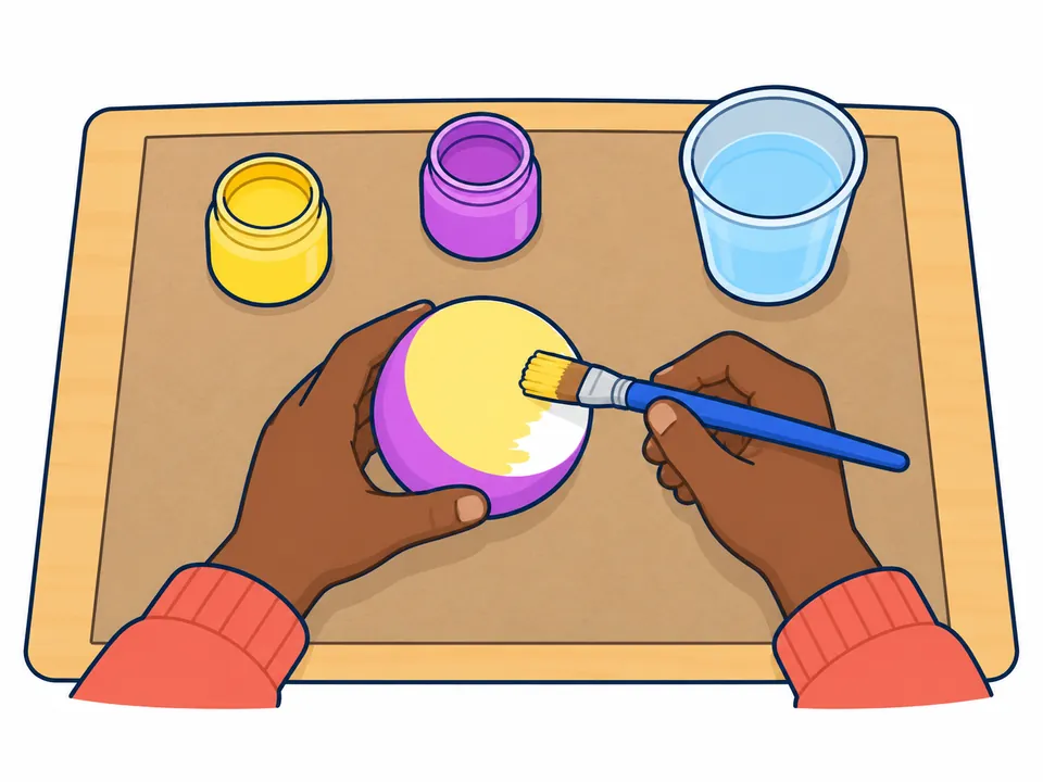 Dos manos sostienen la media bola de unicel con la parte curva ya pintada de morado y pintan de amarillo claro la cara plana con un pincel; en la mesa hay dos botes de pintura y un vaso de agua.