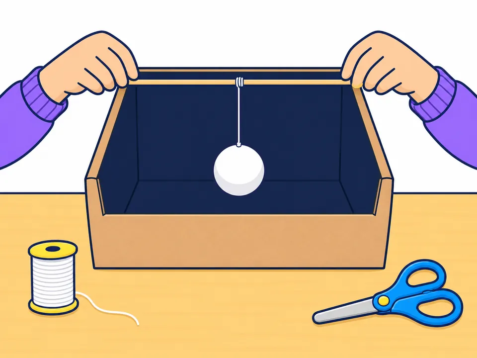 Dos manos colocan un palito de brocheta de lado a lado sobre una caja abierta pintada de negro por dentro; de su centro cuelga con un hilo blanco una bola blanca, en medio de la caja; al lado, un carrete de hilo y unas tijeras.